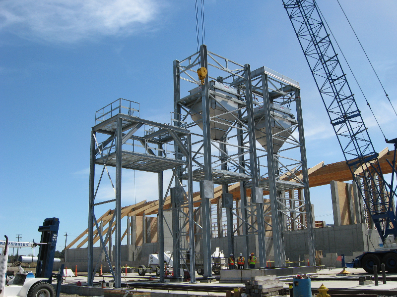 A galvanized structural steel frame being set by crane against a clear sky